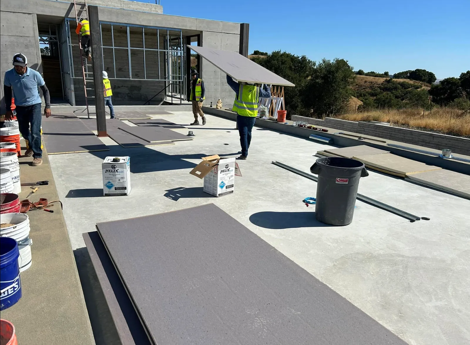 Rea Roofing crew carrying insulation and cover board panels across the flat roof deck of a concrete building under construction, with two-part adhesive pails staged nearby and oak hills beyond