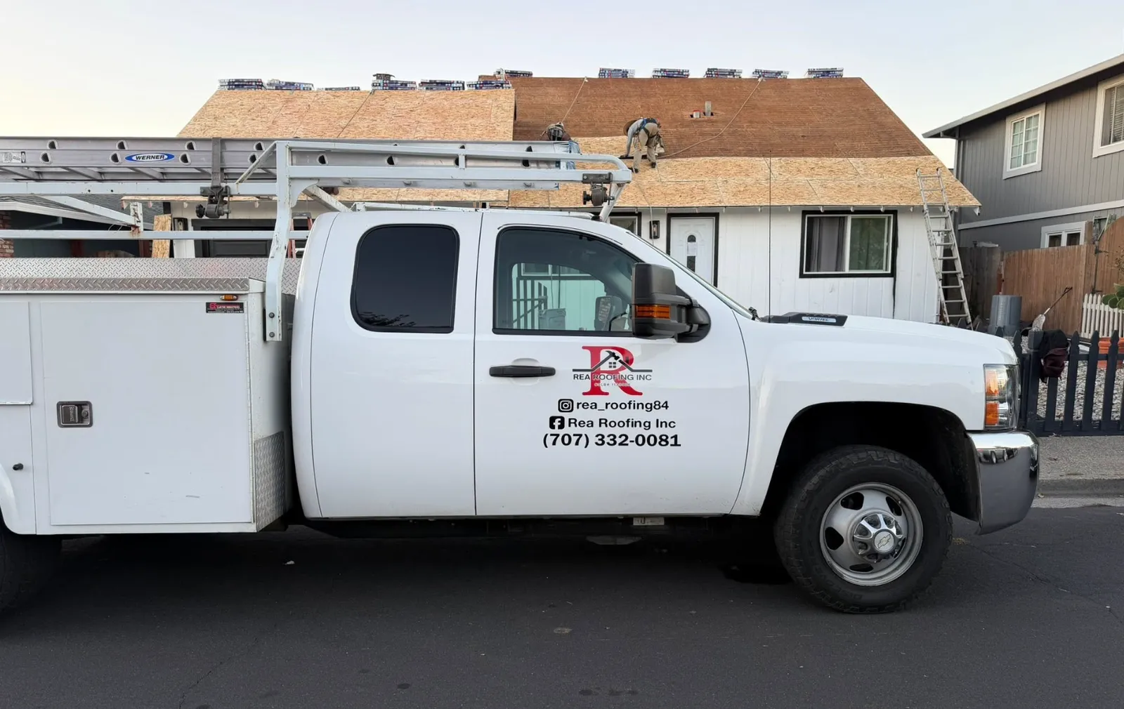 The Rea Roofing Inc. service truck with ladders on the rack parked in front of a single-story home stripped to bare plywood sheathing, with shingle bundles staged along the ridge and a crew member working on the roof