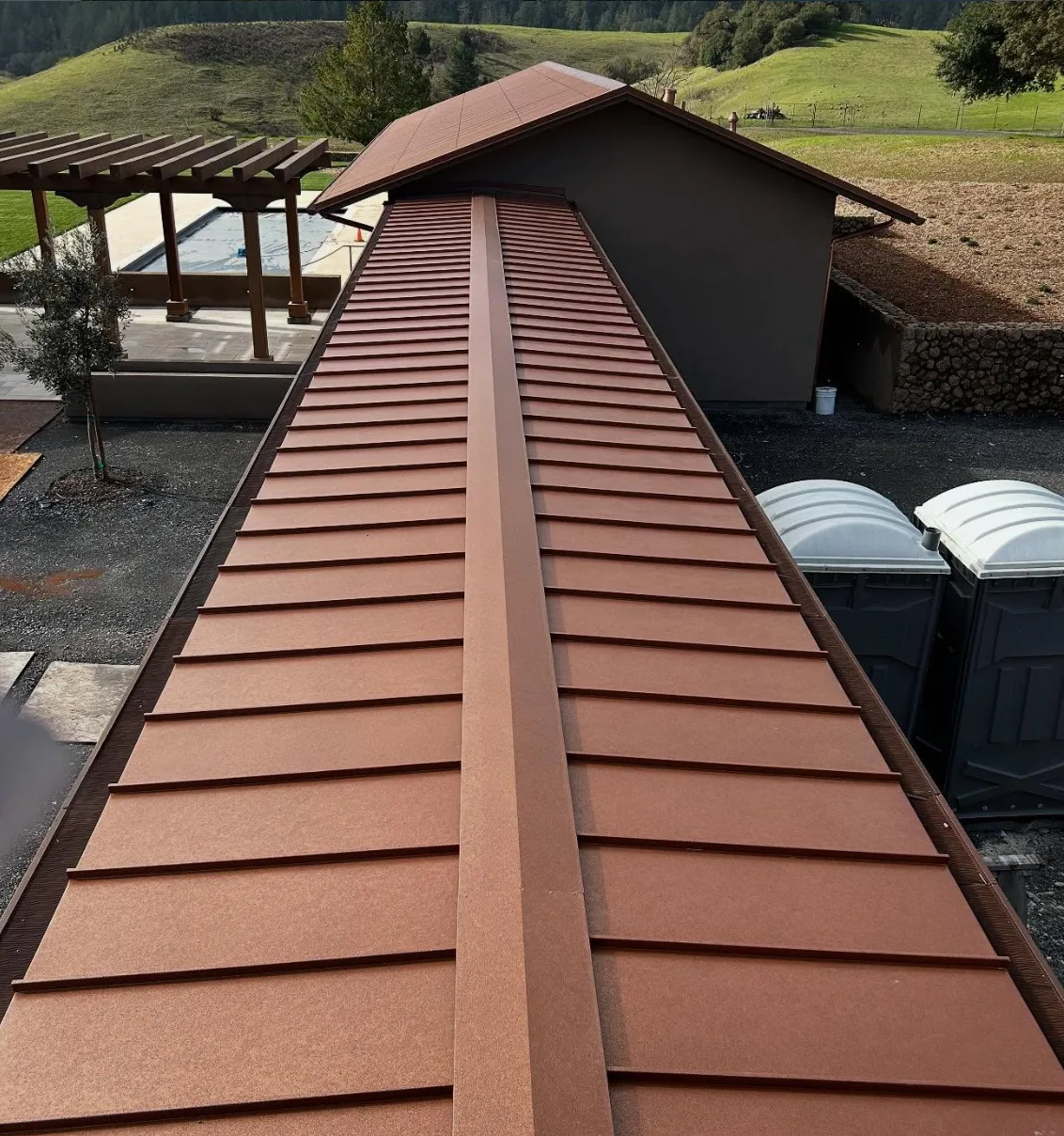 Terracotta standing seam metal roof running the full length of a hillside estate home, with a timber pergola, pool and green North Bay hills behind it